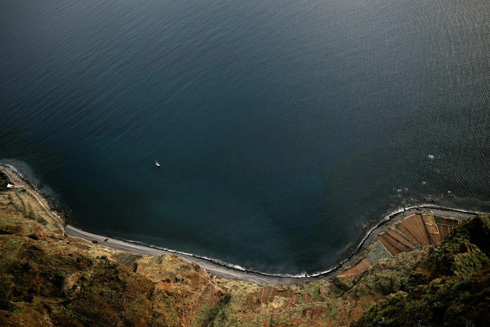 Atlantic coastline with a small sailboat below steep cliffs in Madeira